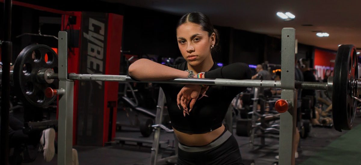 Confident female athlete posing with a barbell in an indoor gym setting.